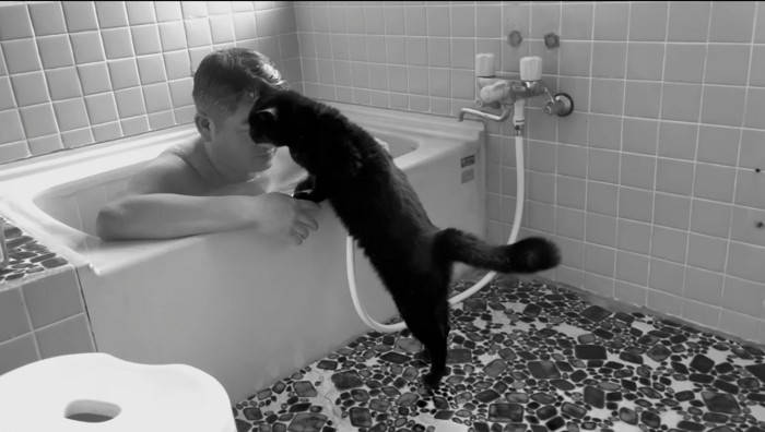 浴槽の縁に前足をかけて、男性の顔に自分の顔を近づける猫