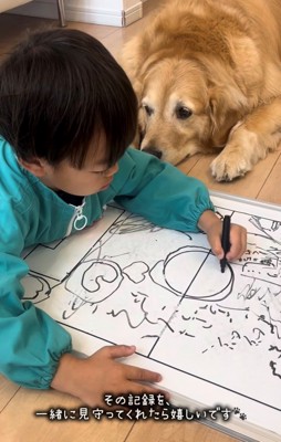 絵を描く子どものそばで伏せて見守る犬