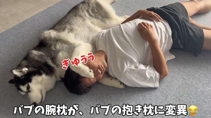 床に寝転がっている男性の頭の下に前足を入れるハスキー犬