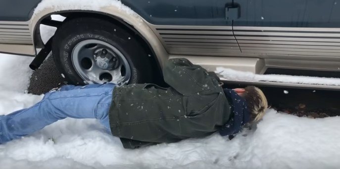 雪の上に寝そべって車の下をのぞく人